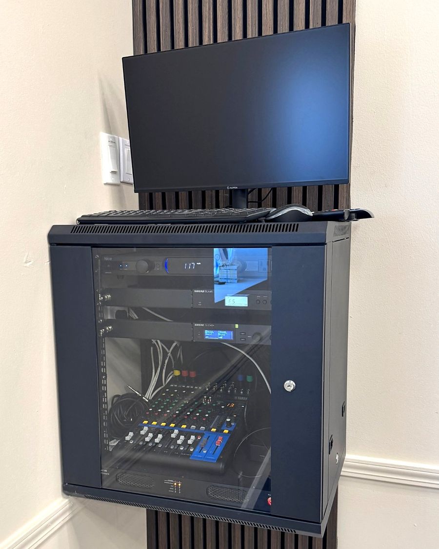 Sound system in a locked, glass-front wall cabinet on a wood slat wall, with wireless microphone receivers, a mixing console and a power amplifier, and a monitor on top